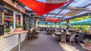 alt="Überdachte Restaurantterrasse mit Holztischen, grauen Stühlen, roten Sonnensegeln und grünen Pflanzen am Rand