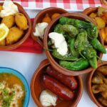alt="Bunte Auswahl spanischer Tapas mit Gambas, Chorizo, Papas Bravas, Pimientos de Padrón und frittiertem Fisch in kleinen Tonschalen.